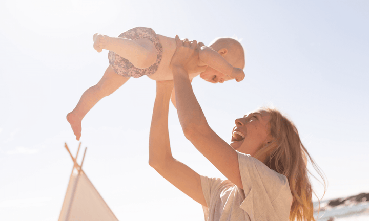 blonde woman lifting small baby wearing a swim nappy into air 